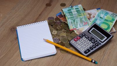 מטבעות ושטרות של כסף ישראלי פזורים ע השולחן ויחד איתם נמצא מחשבון, פנקס ועיפרון כדי לחשב איפה הכי בדאי לחסוך בתכנית חיסכון לכל ילד