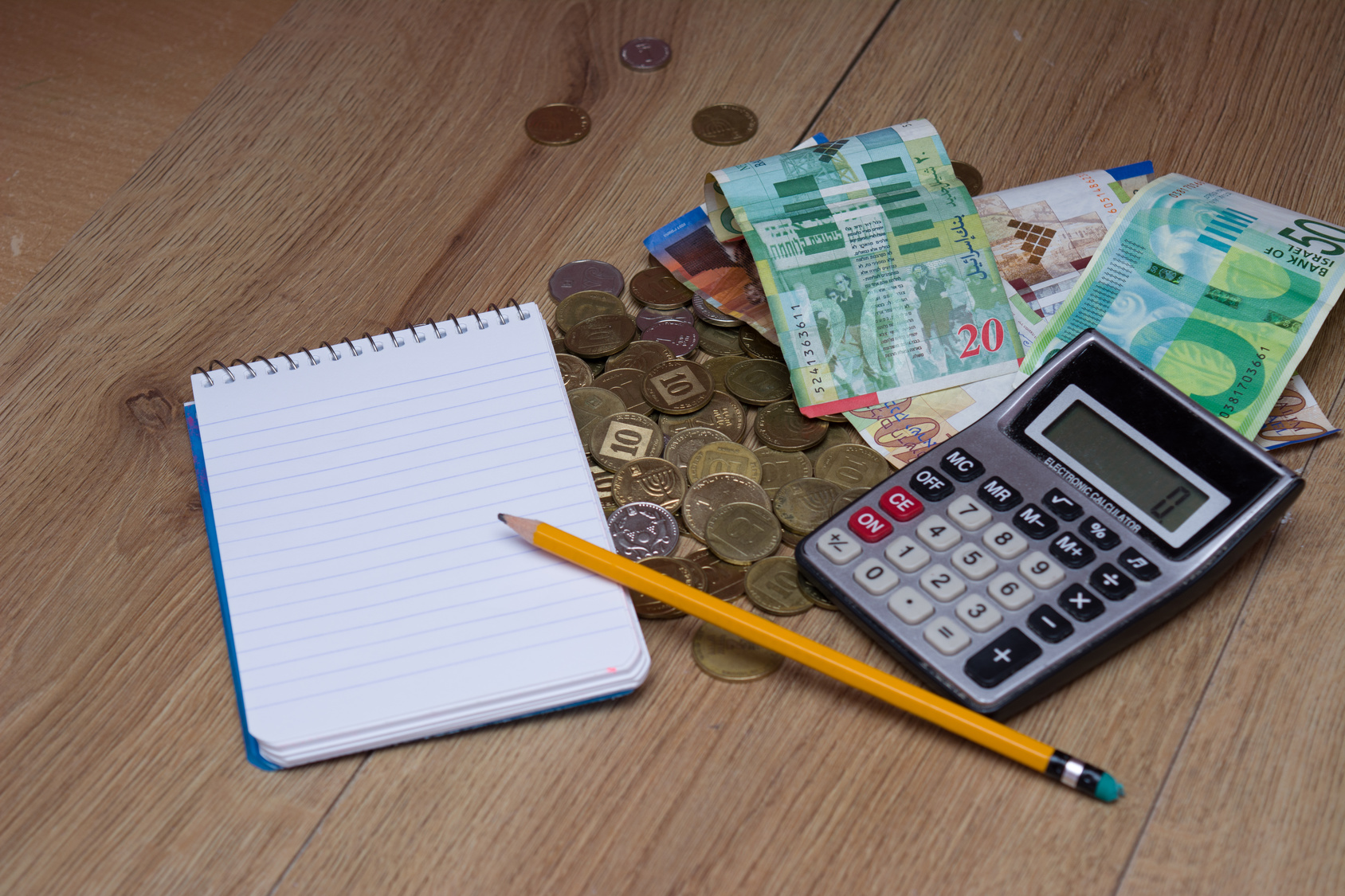 מטבעות ושטרות של כסף ישראלי פזורים ע השולחן ויחד איתם נמצא מחשבון, פנקס ועיפרון כדי לחשב איפה הכי בדאי לחסוך בתכנית חיסכון לכל ילד