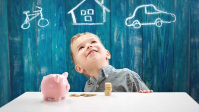ילד בן שלוש יושב על השולחן עם כסף וקופת חיסכון בצורת חזירון. ילד שמח עם מטבעות. ילד שחולם על הבית ועל רכב ואופניים